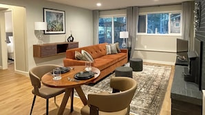 A modern living room with a sofa, two chairs, a dining table with plates and glasses, a fireplace, and a city view through the window. - Walk to Beach + Village: Bainbridge Island Gem! (Bainbridge Island)