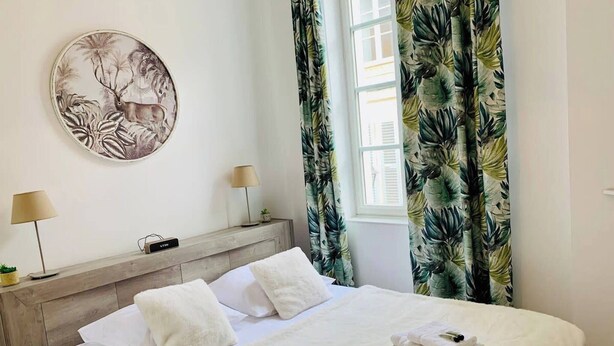 A hotel room with a bed, two bedside lamps, a VTR device, a wall-mounted painting, and tropical curtains. - Le Grand Cloutiers, Centre Ville, LR135 (La Rochelle)
