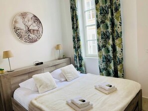 Ein Hotelzimmer mit einem Bett, zwei Nachttischlampen, einem Video-Rekorder, einem Wandbild und tropischen Vorhängen.