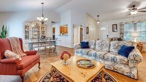 A spacious living room with a red armchair, a floral sofa, and a wooden coffee table. A dining area with a chandelier and a kitchen with wooden cabinets is visible in the background. - 2 Mi to Ksu: Family Home w/ Games, Near Parks (Kennesaw)