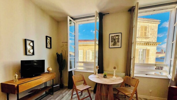 A room with a wooden floor, a TV on a wooden stand, a round table with chairs, and large windows with a view of buildings. - Le Petit Marché, Hypercentre, LR125 (La Rochelle)
