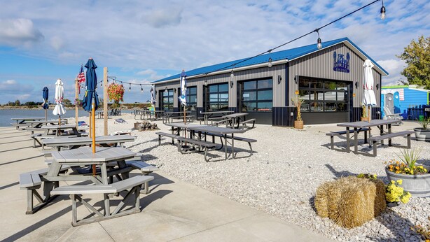 An outdoor seating area with picnic tables and umbrellas, a building with a blue roof, and a view of the water. - Walk to Lagoon Saloon! Chic Home on Nugent's Canal (Port Clinton)