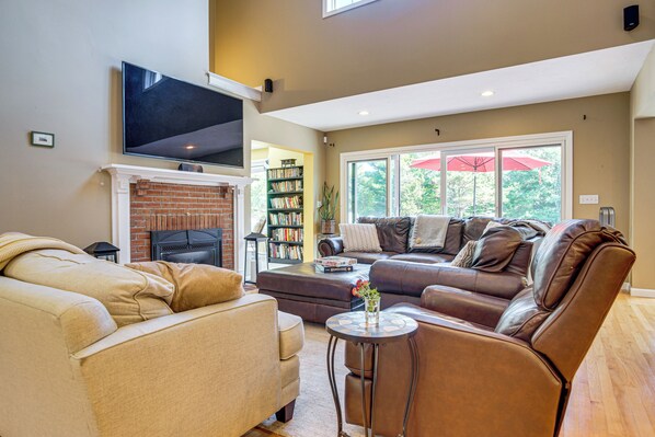 A spacious living room with a fireplace, leather sofas, a coffee table, and a bookshelf. - 3 Mi to Megansett Beach: Home w/ Pool & Game Room (East Falmouth)