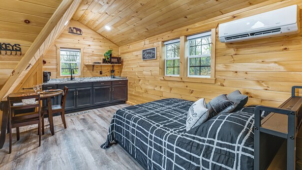 L’intérieur chaleureux d’un chalet avec un décor en bois, un lit recouvert d’une couverture à carreaux noirs et blancs, une table à manger avec des chaises, un coin cuisine et une fenêtre donnant sur les arbres.