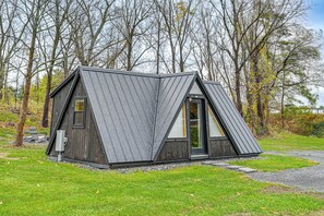 A small, triangular wooden cabin with a sloped roof, surrounded by trees and a grassy area.