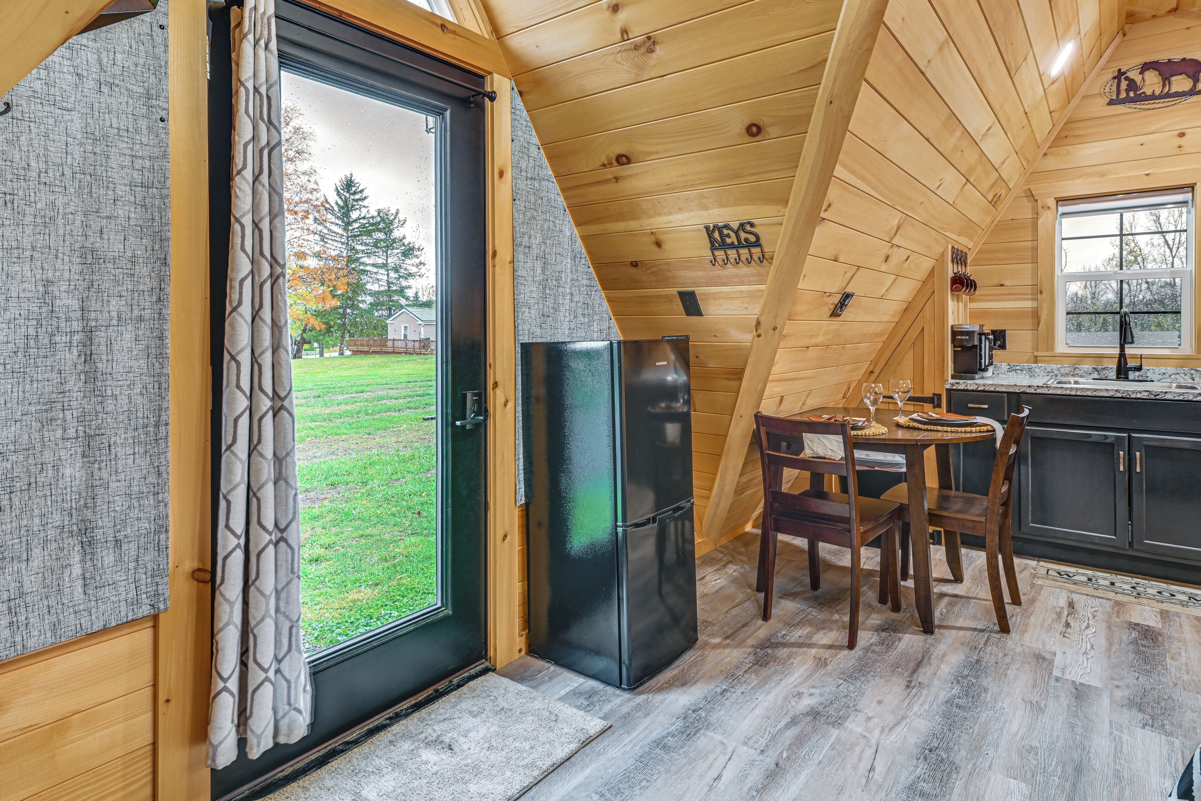 A wooden cabin interior with a dining area, a black refrigerator, and a view of the outdoors.