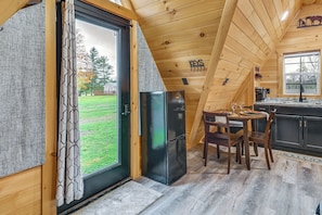 A wooden cabin interior with a dining area, a black refrigerator, and a view of the outdoors.
