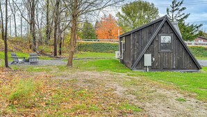 Une petite cabane en bois avec un toit triangulaire, une fenêtre et une porte. On y trouve également deux chaises Adirondack et une table de pique-nique à proximité.