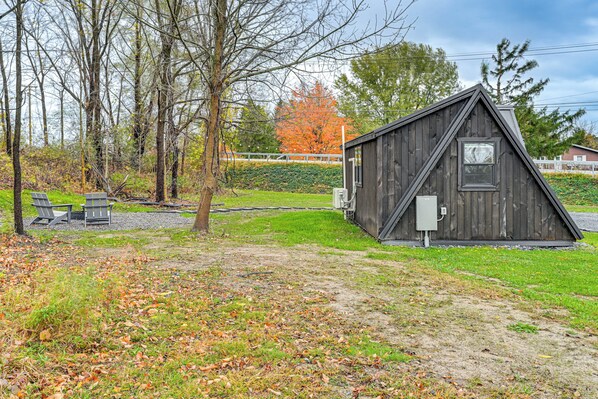 A small wooden cabin with a triangular roof, a window, and a door. There are two Adirondack chairs and a picnic table nearby.