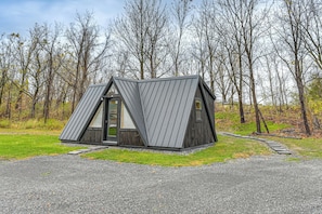 A modern, triangular-shaped cabin with a metal roof, surrounded by a gravel driveway and a grassy area with trees in the background. - 5 Mi to Wine Trails: Finger Lakes Tiny Home (Ovid)