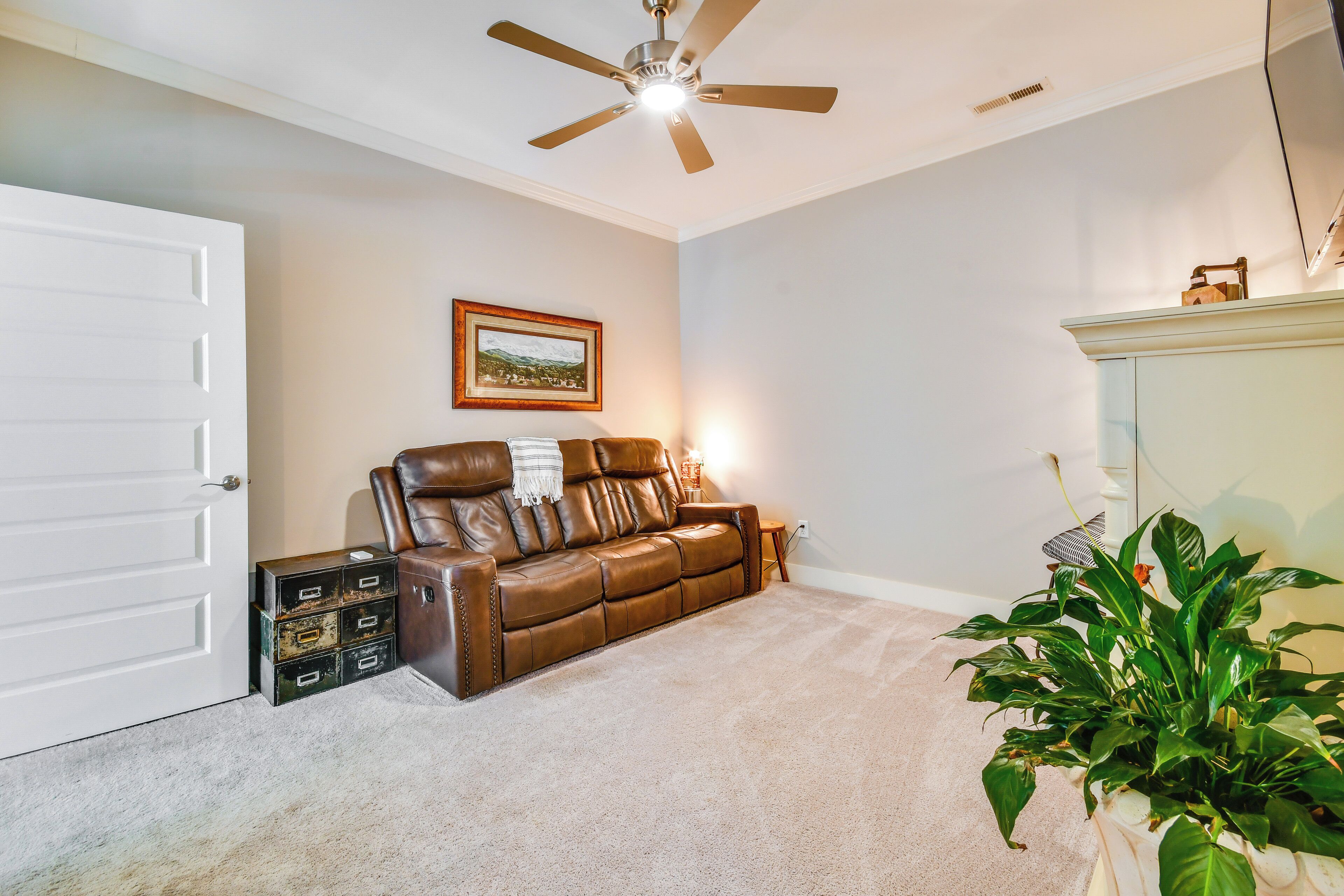 A living room with a brown leather sofa, a ceiling fan, a framed picture on the wall, and a potted plant.