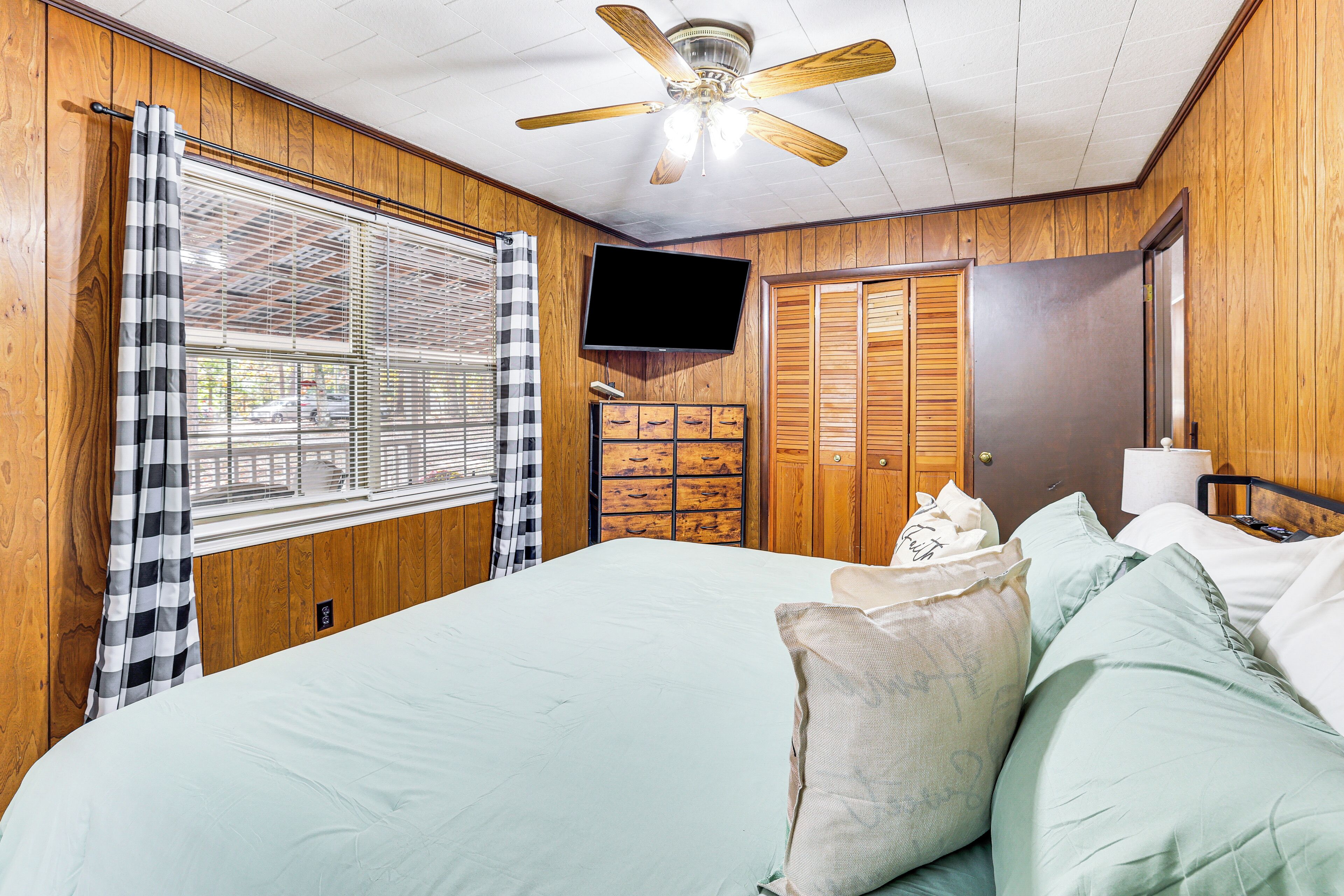 A bedroom with a bed, a ceiling fan, a TV, wooden walls, a window with blinds, and a door with a mirror.