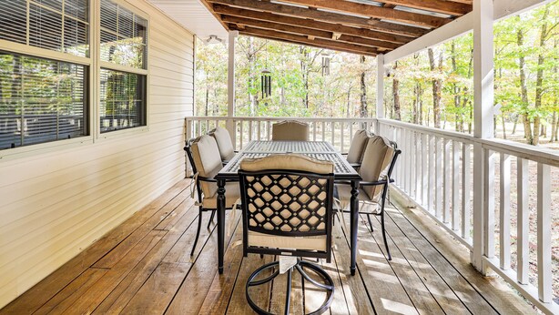 A covered patio with a wooden table and chairs, surrounded by white railings and large windows.