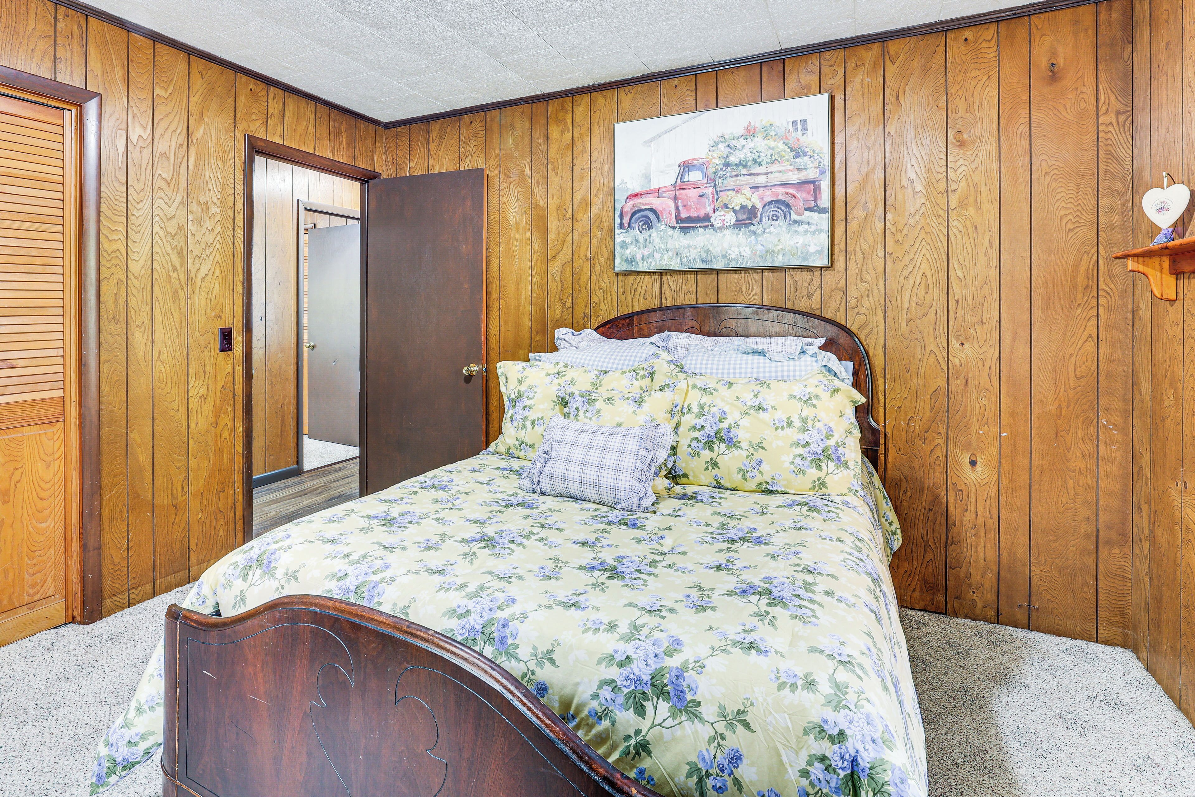 A bedroom with wooden paneling, a bed with floral bedding, a door, and a painting of a red truck.