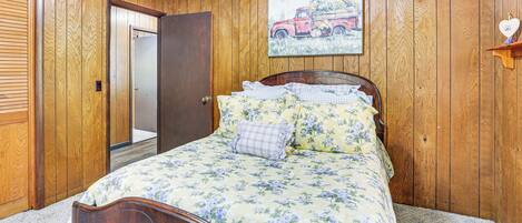 A bedroom with wooden paneling, a bed with floral bedding, a door, and a painting of a red truck.