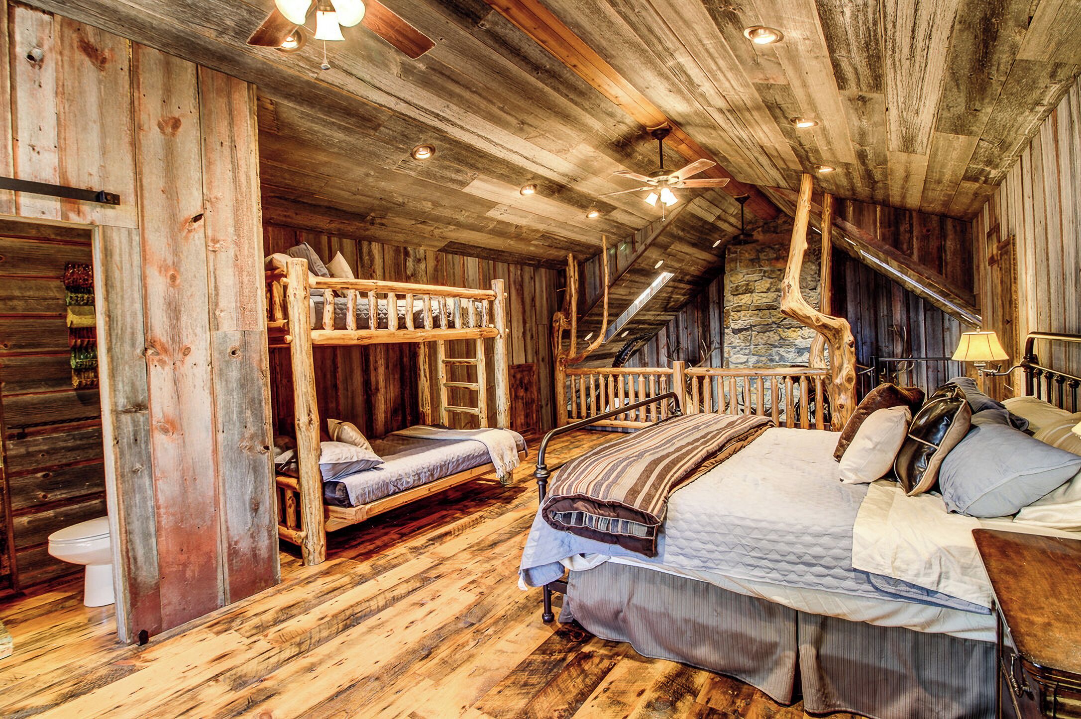 A rustic wooden room with a bunk bed, a single bed, a ladder, and a stone wall.