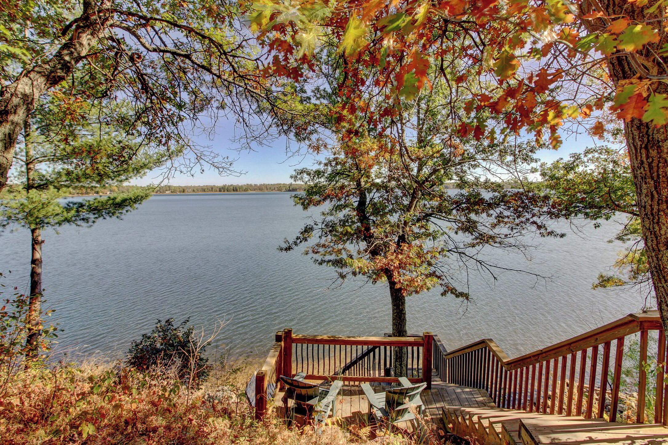 Eine Holzterrasse mit Stühlen, die auf einen ruhigen See mit Herbstlaub blickt, umgeben von Bäumen.