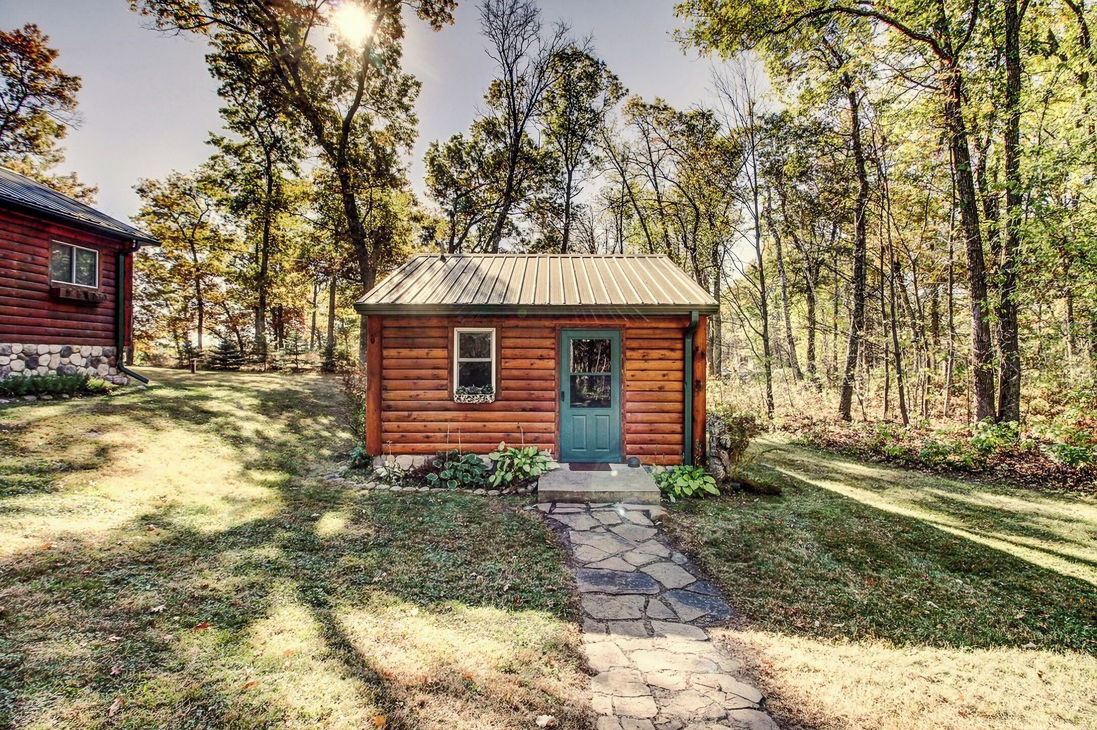 A small wooden cabin with a green door and windows, surrounded by a stone pathway and a grassy area, set in a forested landscape.