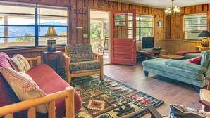 Ein gemütliches Zimmer mit Holzvertäfelung, einer roten Tür, einem grünen Sofa, einem gemusterten Sessel und einem großen Fenster mit Blick auf die Berge.