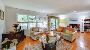 A living room with a sofa, armchair, and a glass coffee table. There are windows with curtains, a bookshelf, and a lamp.
