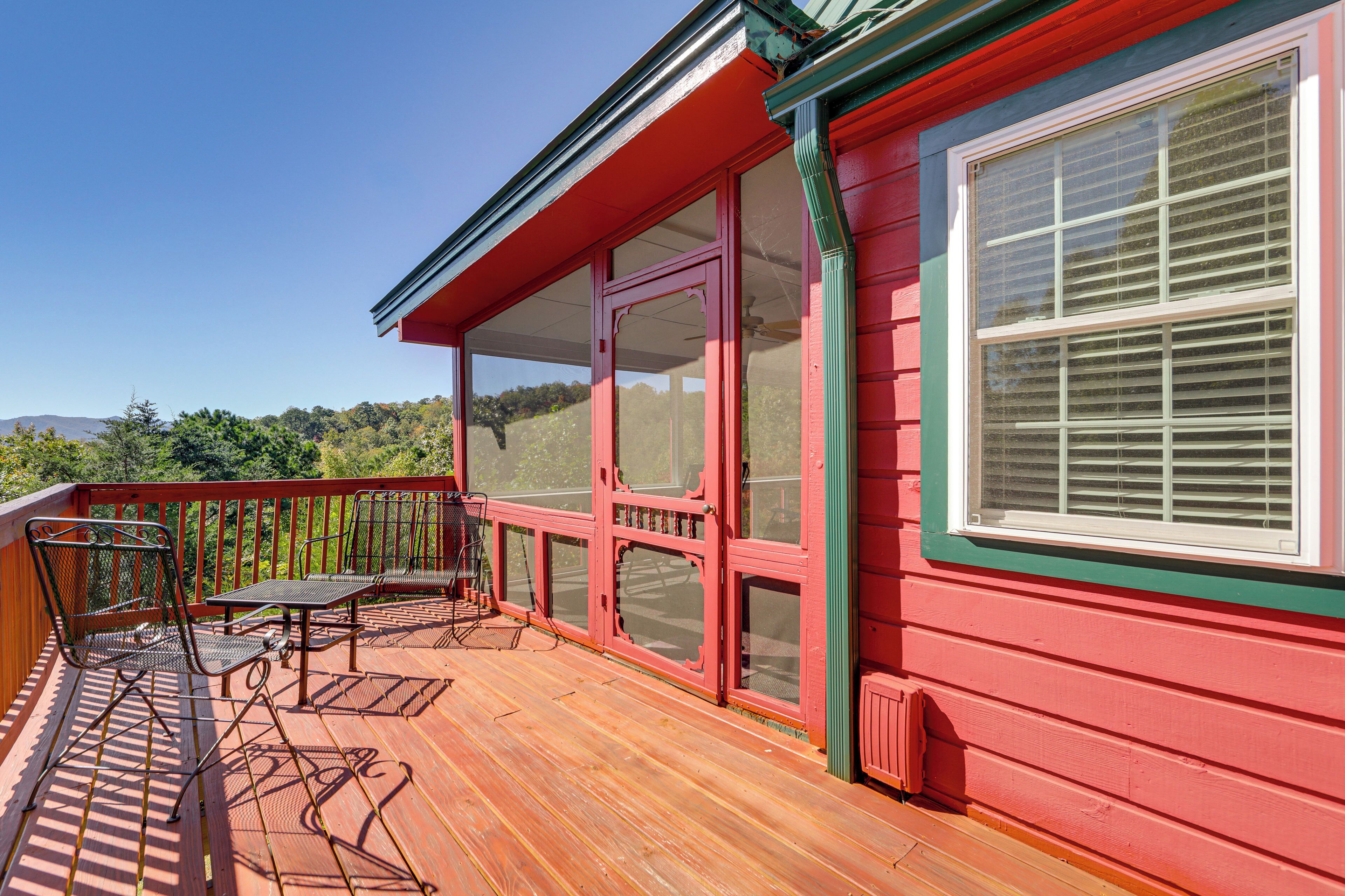 A wooden deck with metal chairs and a table, a red building with a large window, and a view of trees and hills in the background.