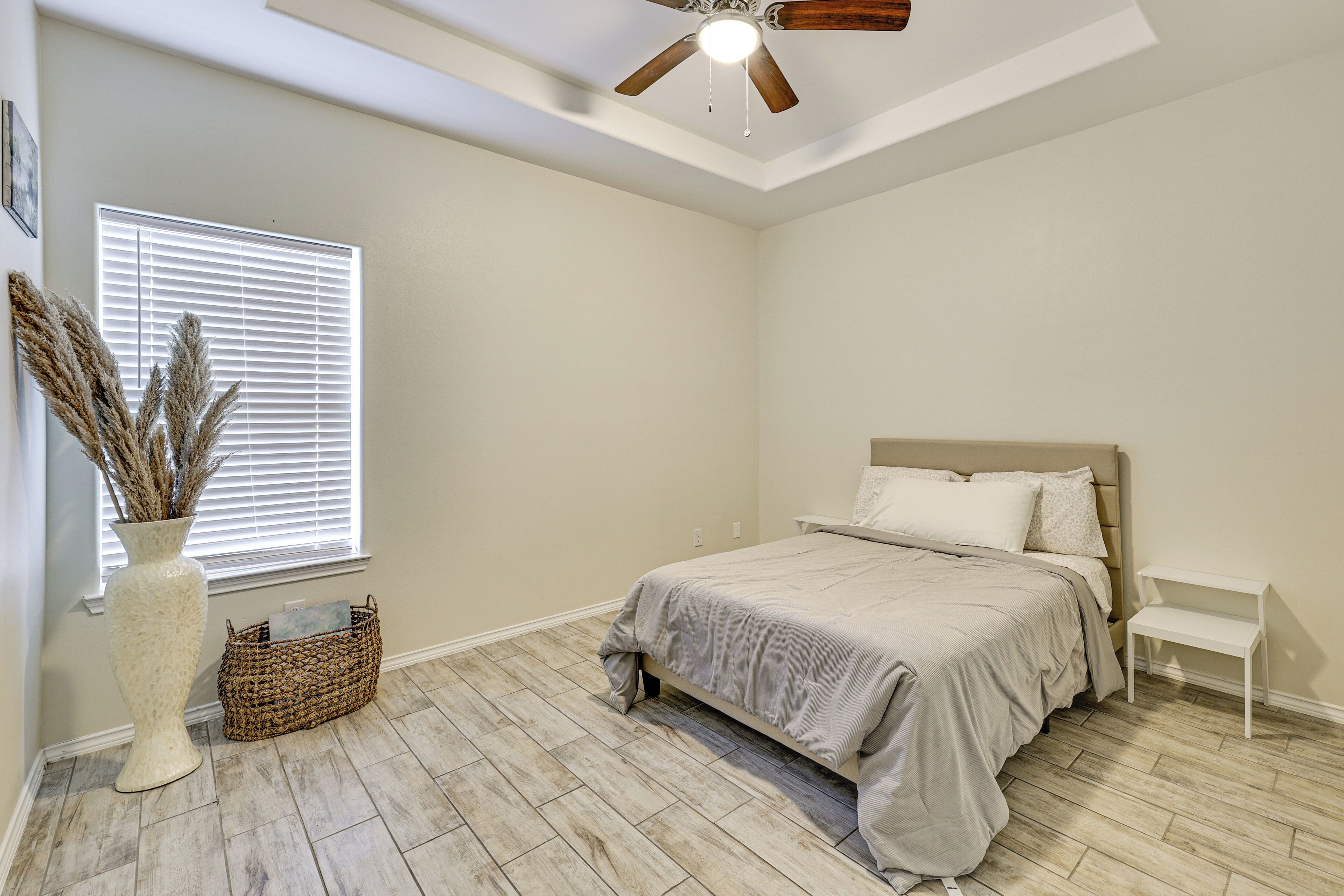 A bedroom with a bed, a ceiling fan, a window with blinds, a vase with dried plants, and a wicker basket.