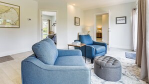 A modern living room with a blue sofa, a grey ottoman, and a blue armchair. There is a small table with a lamp and a book. The room has a large window with curtains, a map on the wall, and a 'HAPPY' sign. - Boat Dock & Yard: Waterfront Okeechobee Retreat (Okeechobee)