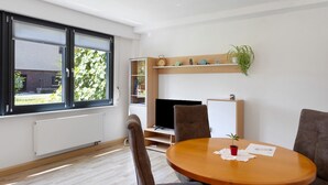 A modern living room with a large window, a flat-screen TV, a wooden dining table, and a built-in shelf with plants. - Apartment 'Vollmarshausen' with Shared Garden and Wi-Fi (Lohfelden)
