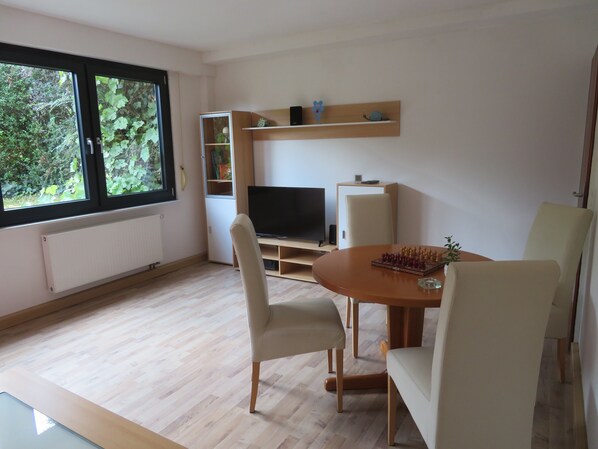 A modern living room with a TV, wooden shelves, a dining table, and chairs.