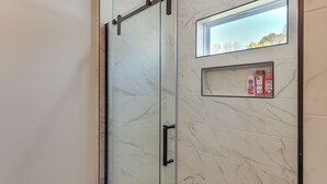 Eine gläserne Dusche mit Marmorfliesen, einem kleinen Ablagebereich für Toilettenartikel und einem Fenster darüber.