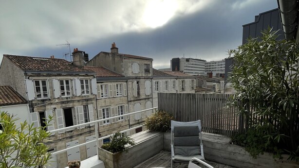 A wooden deck with a round table and two chairs, surrounded by potted plants and overlooking a cityscape. - Le bonpland, centre ville, LR122 (La Rochelle)