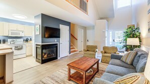 A modern kitchen with white cabinets, a microwave, and a fireplace. There is a dining area with a wooden table and a sofa. A staircase leads to an upper level.