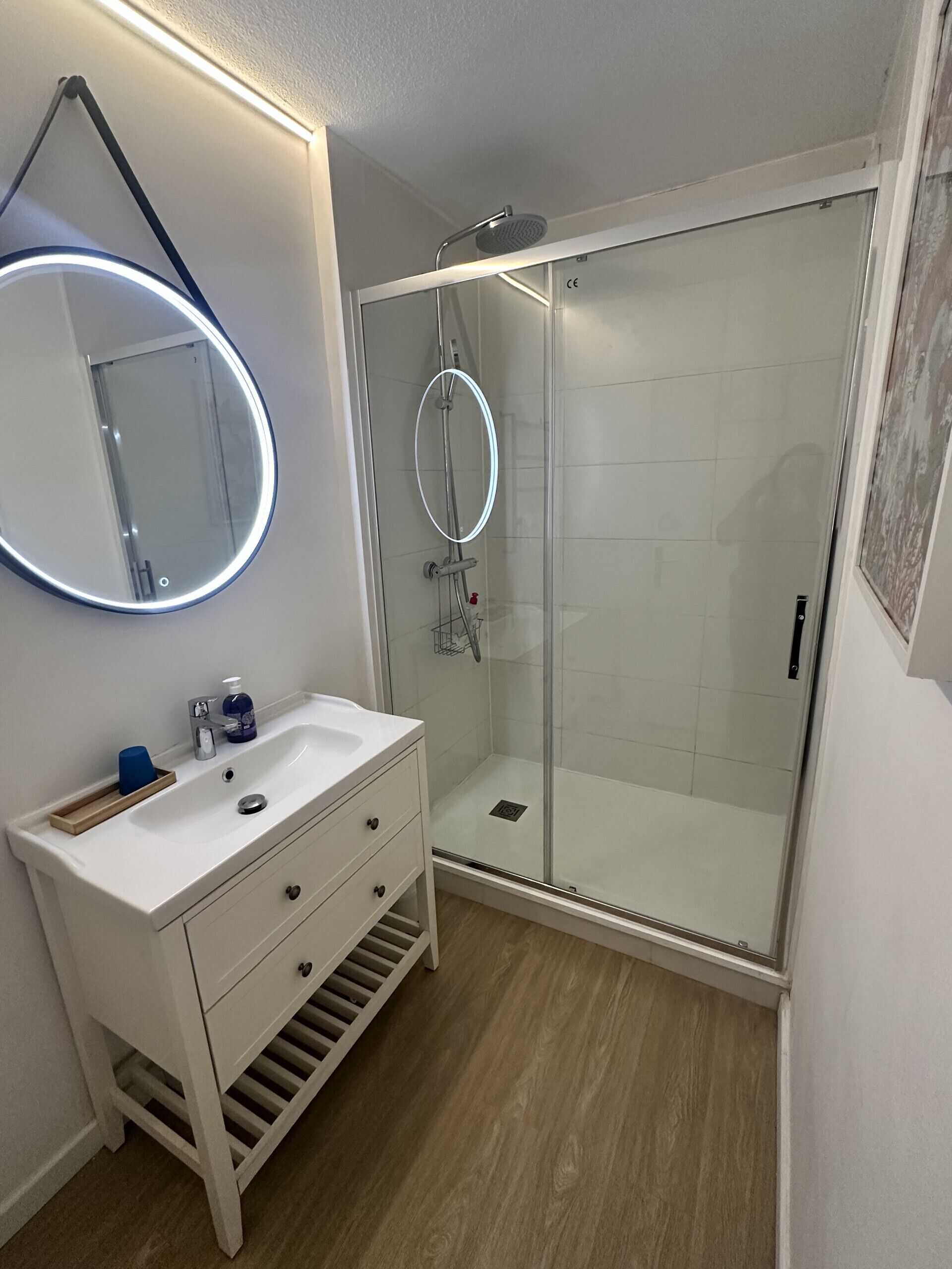 Un bany modern amb dutxa amb envàs de vidre, un lavabo blanc amb mirall i un armari de fusta.