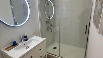 Un bany modern amb dutxa amb envàs de vidre, un lavabo blanc amb mirall i un armari de fusta.