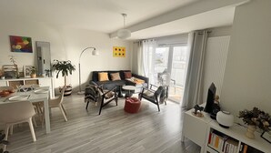 Un salon moderne avec un canapé, une table basse et un coin repas. On y trouve une bibliothèque remplie de livres et une plante en pot.