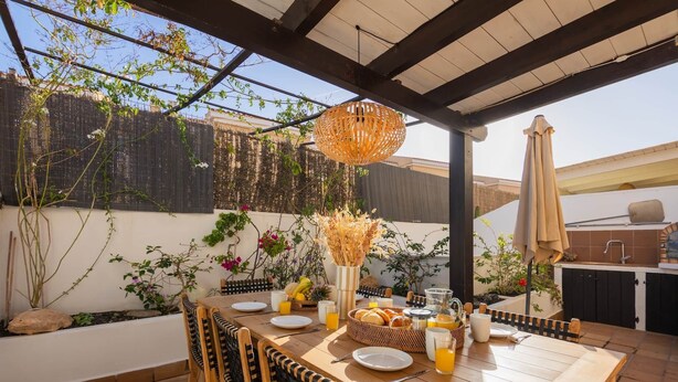 Eine Terrasse im Freien mit einem Holztisch, gedeckt für ein Frühstück, umgeben von Korbstühlen, einer hängenden Leuchte und verschiedenen Pflanzen.