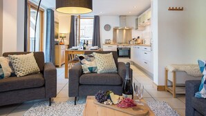 A modern living room with a sofa, a coffee table with food and drinks, and a kitchen in the background. - More Mountain - Apartment Central in the heart of Morzine with Outdoor Jacuzzi. Less than 5 minutes walk to ski lifts & bars. (Morzine)