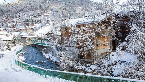 A snow-covered village with a river, wooden houses, and a bridge. - More Mountain - Apartment Central in the heart of Morzine with Outdoor Jacuzzi. Less than 5 minutes walk to ski lifts & bars. (Morzine)