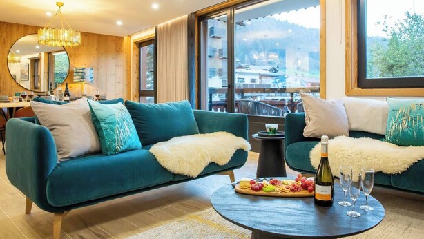 A modern living room with a teal sofa, a round coffee table with a bottle and glasses, and a view of a mountainous area through large windows. - More Mountain  - Demi Lune. Central Morzine location and less than 2 minutes walk to main ski lifts, bars and restaurants. (Morzine)
