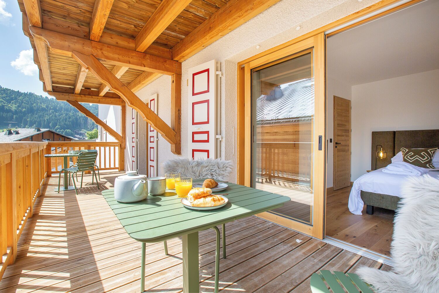 Une terrasse en bois avec une table préparée pour le petit-déjeuner, un lit recouvert d’une couette blanche et une vue sur les montagnes.