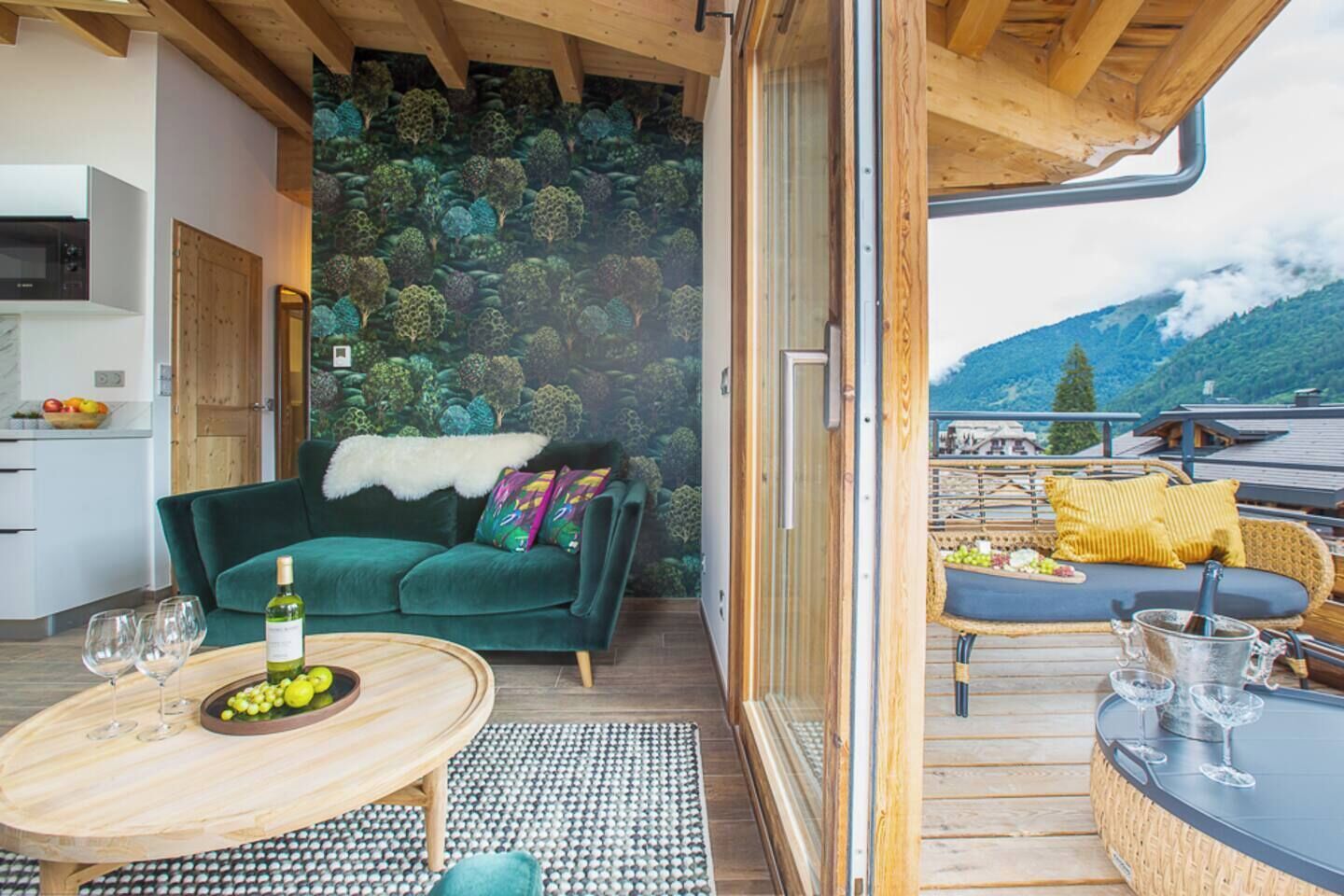 A modern living space with a green sofa, a wooden table with wine glasses and grapes, and a balcony with a view of mountains.