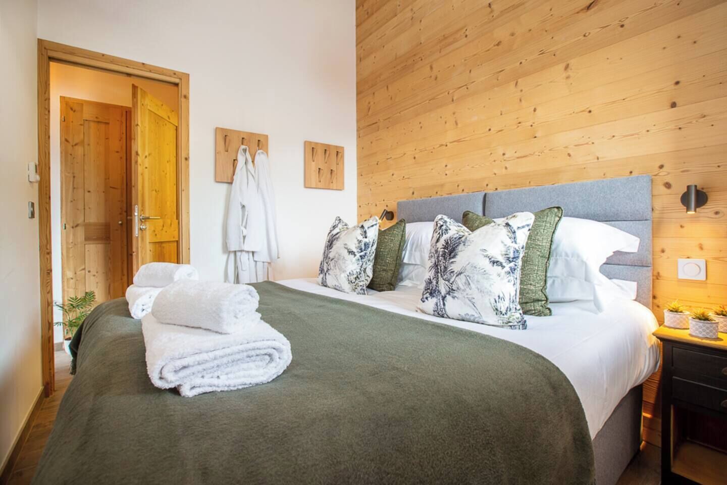 A neatly made bed with a green bedspread and white towels, wooden paneling on the walls, and a wooden door.