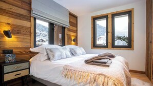 Un dormitorio con pared de madera, una cama con ropa blanca, dos ventanas y una mesita de noche con lámpara.