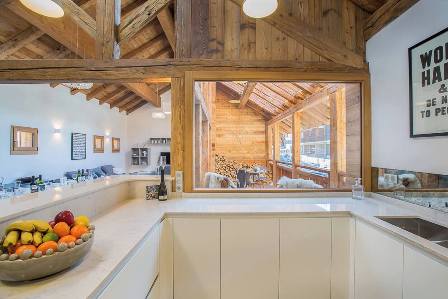 Une cuisine moderne dotée d’un grand îlot central, de poutres en bois et offrant une vue sur un espace extérieur rustique.
