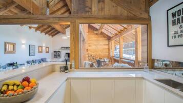 A modern kitchen with a large island, wooden beams, and a view of a rustic outdoor area.