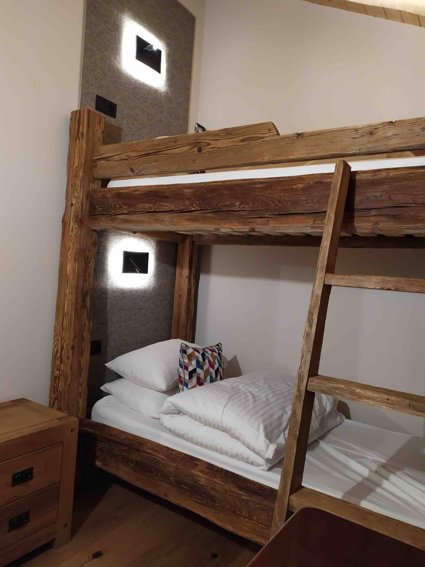 A bunk bed with a wooden frame, a headboard with a textured wall, and a bedside table with drawers.