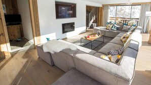 A modern living room with a large sectional sofa, a flat-screen TV, a fireplace, and a staircase leading to an upper level. - More Mountain - Chalet La Godille. Luxury Chalet with large Terrace and Outdoor Jacuzzi split over 3 levels next to the Super Morzine Telecabine. (Morzine)