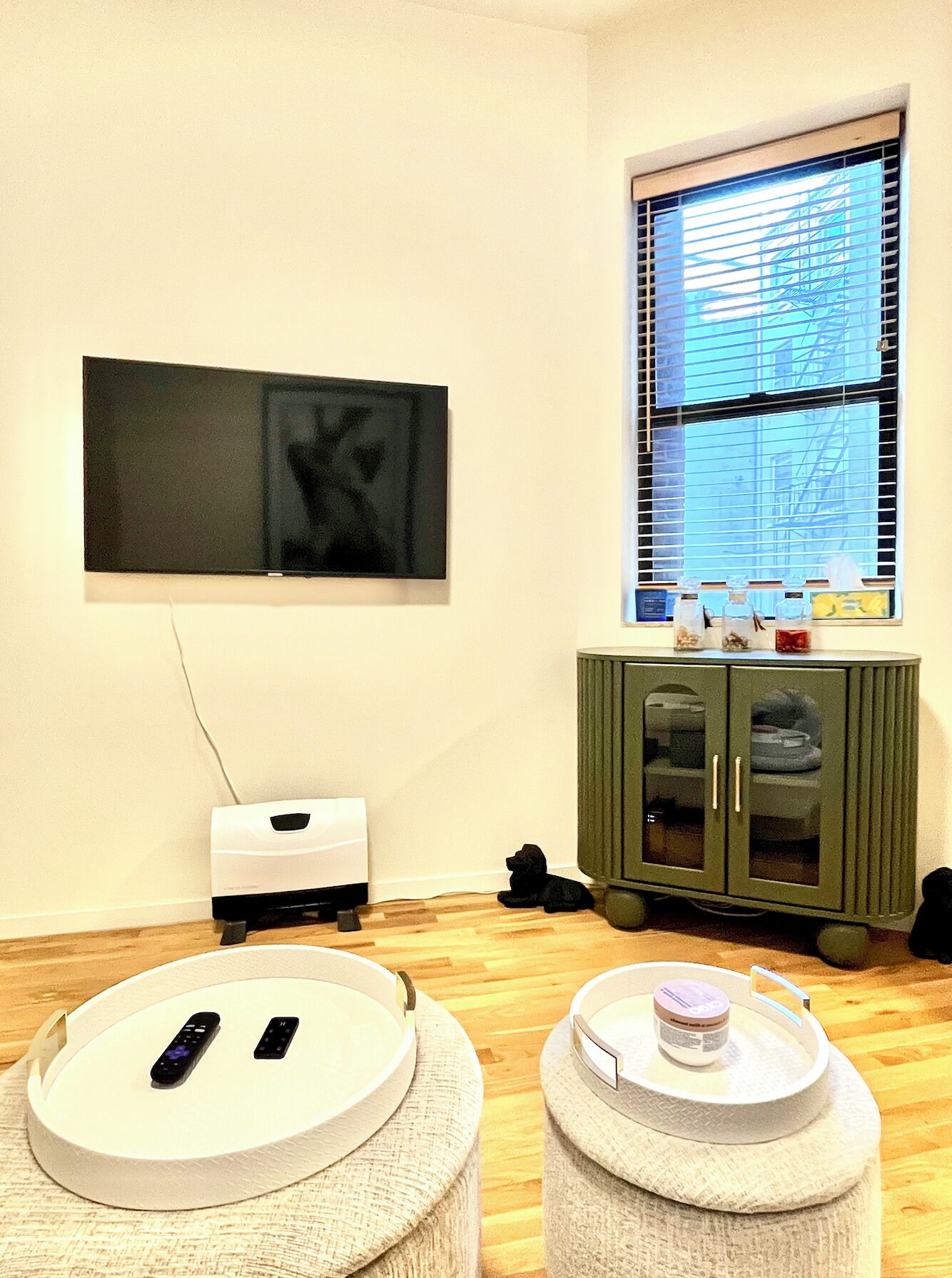 A room with a flat-screen TV mounted on the wall, a window with blinds, a green cabinet, and a small round ottoman with a remote control.
