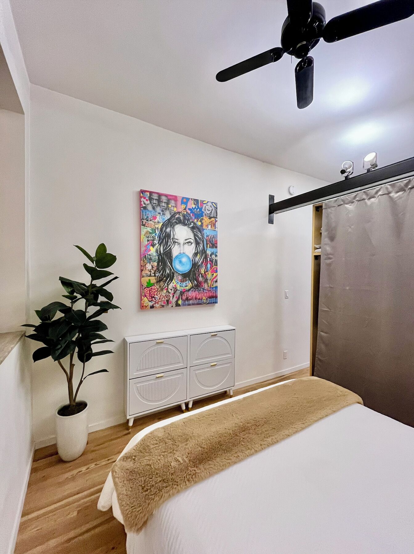 Een slaapkamer met een bed, een plafondventilator, een radiator, een kamerplant en een wanddecoratie.