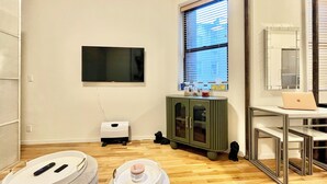 Une pièce dotée d’un téléviseur à écran plat fixé au mur, d’une fenêtre avec des stores, d’un meuble vert, d’une petite table avec un ordinateur portable et d’un pouf blanc avec un récipient.